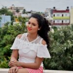 A cheerful woman sitting outdoors in a white lace top against a cityscape backdrop.